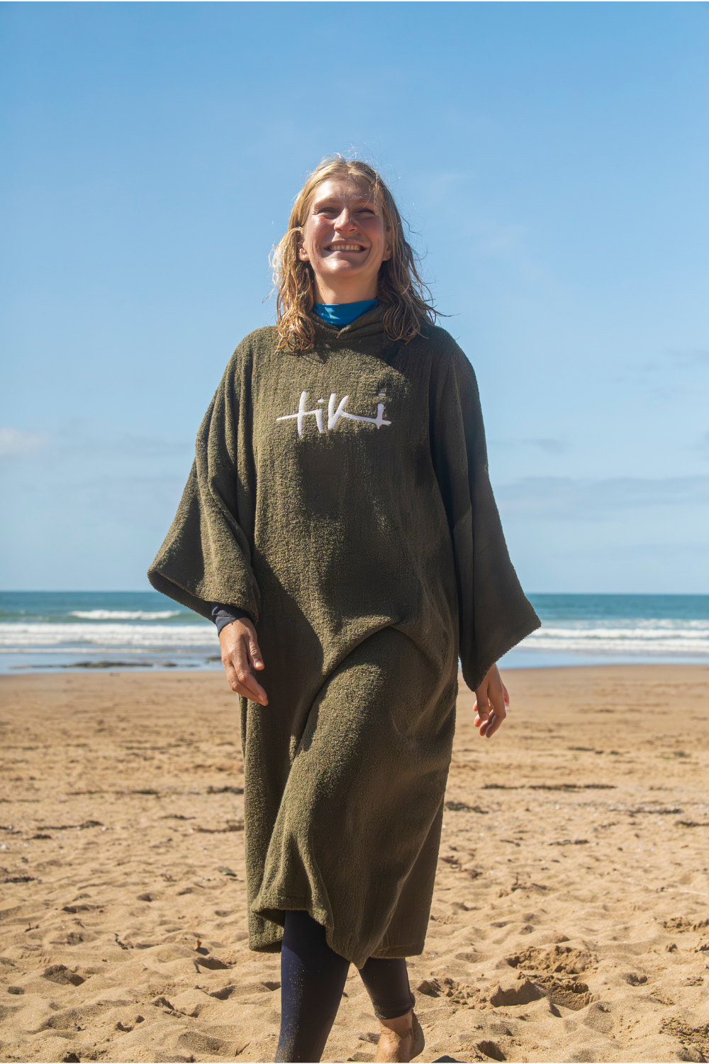 A woman walking on a sandy beach wearing a loose, oversized olive green towelling robe with a white embroidered'Tiki' logo on the chest.