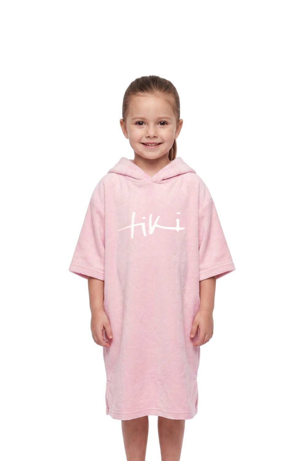 A young girl stands against a white background wearing a light pink hooded towelling robe with the brand name 'Tiki' printed in white across the chest.