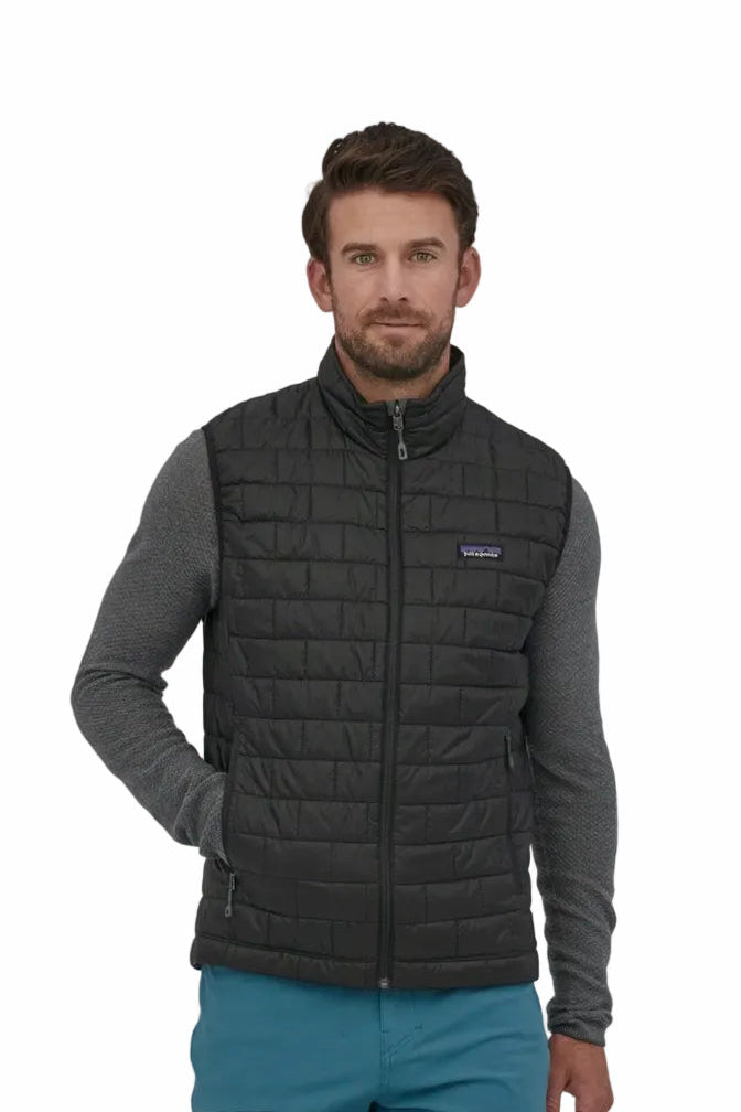 A man wearing a black Patagonia quilted puffer vest over a gray long-sleeve shirt and teal pants, standing against a plain white background.