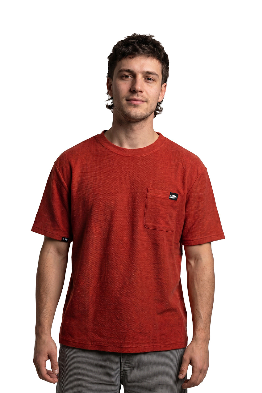Man wearing a terracotta coloured pocket t-shirt with a black logo badge on the pocket and the right sleeve.