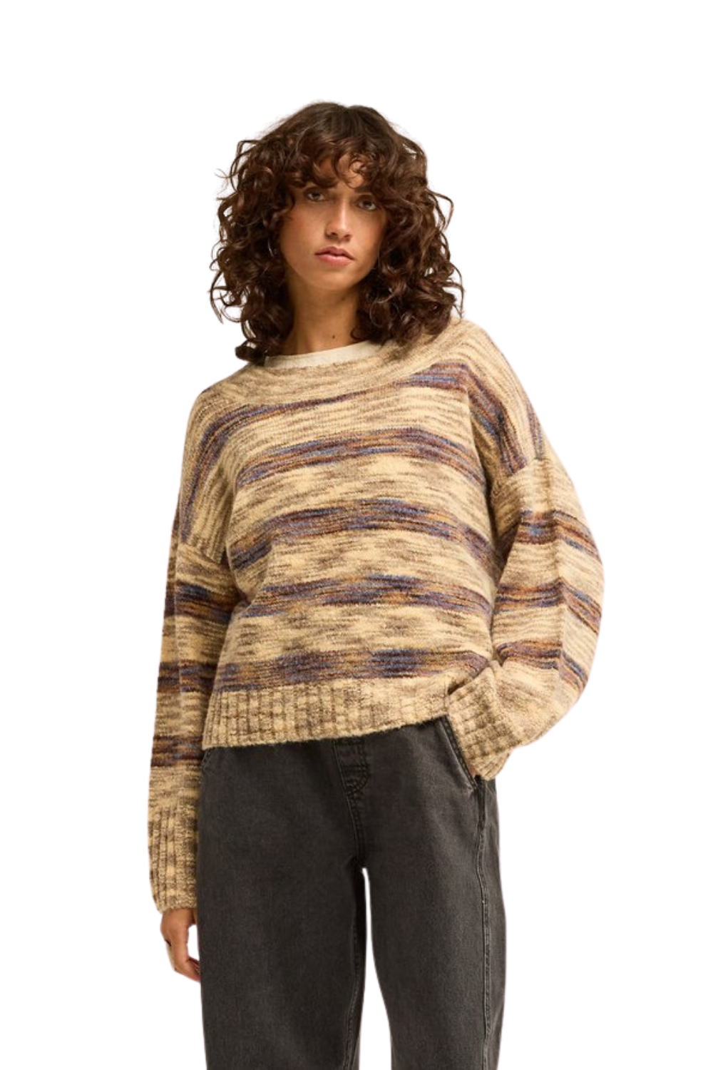 A woman wearing a loose, textured knit sweater with horizontal marled stripes in beige, brown, and muted blue, paired with dark gray jeans, against a plain white background.
