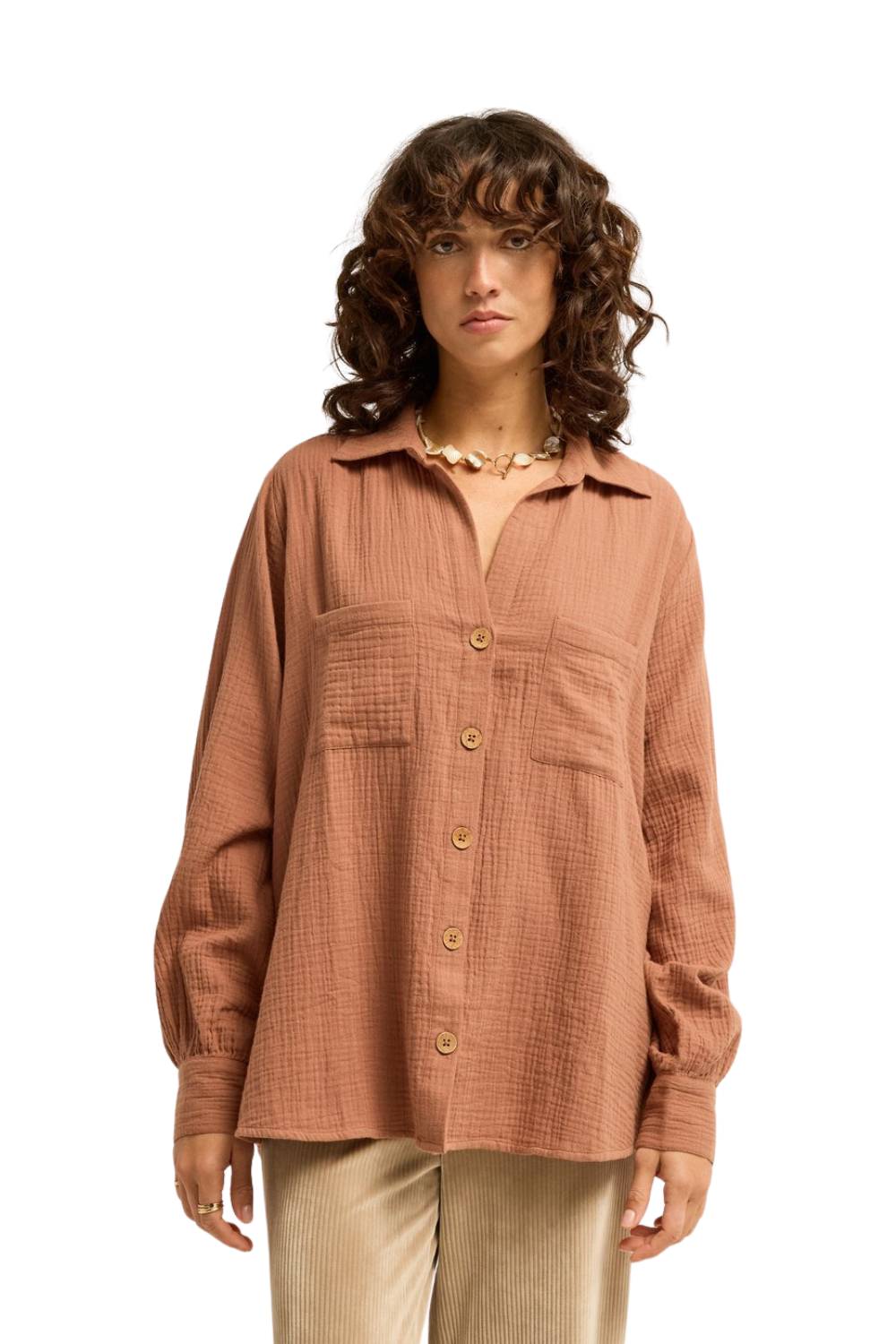 A woman wearing a textured, long-sleeve button-up shirt in a dusty terracotta colour, paired with beige pants, photographed against a solid white background.