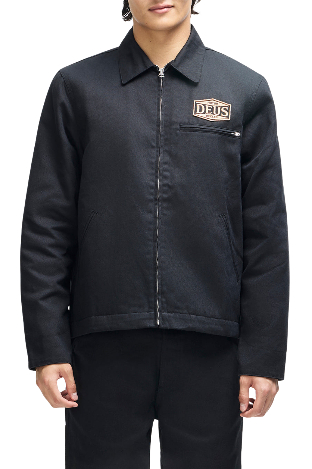 A model wearing a black zip-up jacket with a collar and a 'DEUS' logo patch on the chest pocket, paired with black pants against a white background.