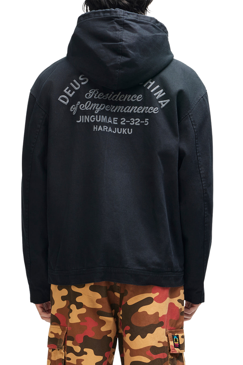 A person shown from the back wearing a black hooded jacket with white printed text on the back reading 'DEUS EX MACHINA' and 'Resilience of Impermanence JINGUMAE 2-32-5 HARAJUKU'. The person is also wearing camouflage cargo pants in tan, brown, and red tones against a plain white background.