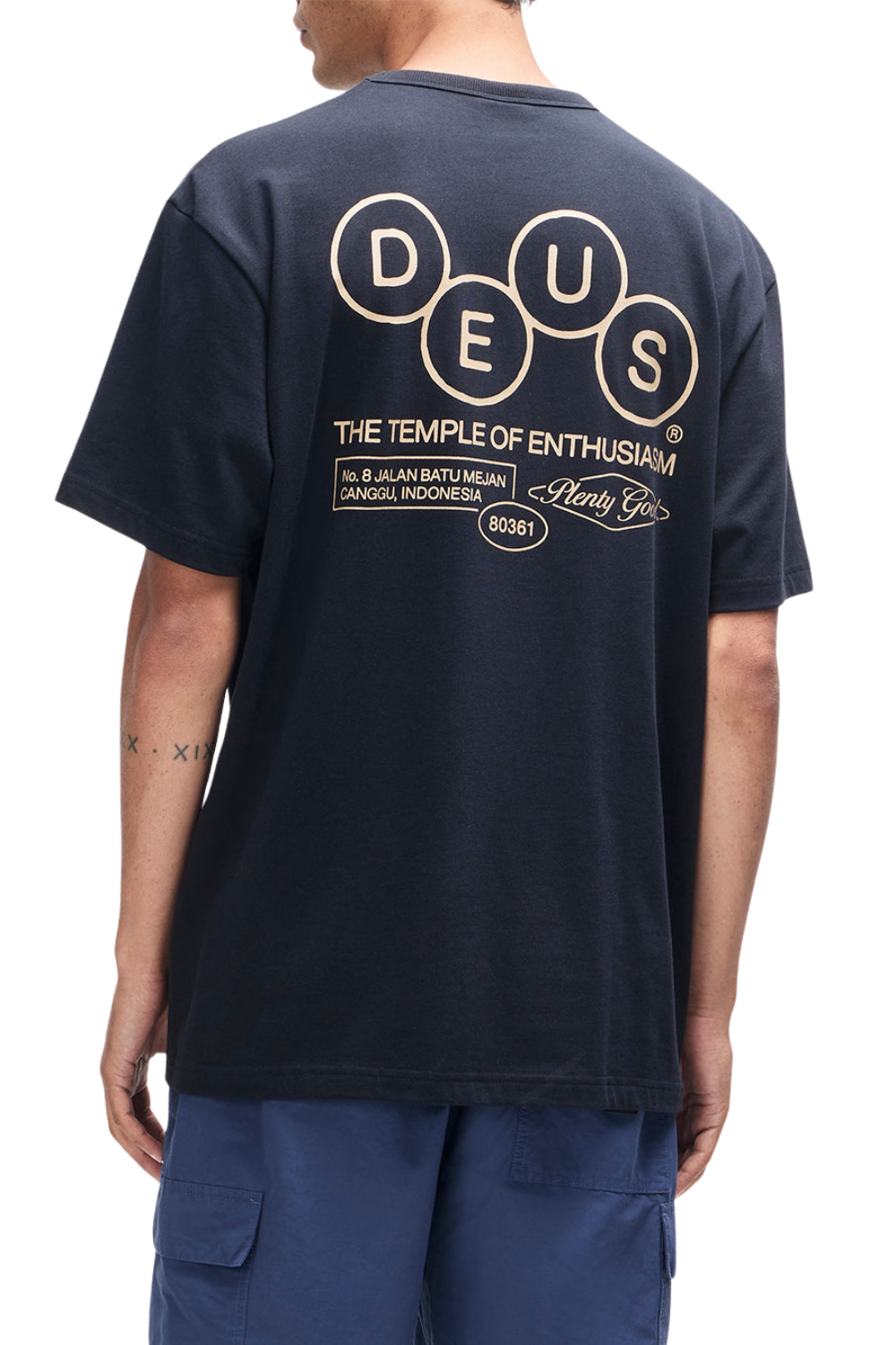 Person wearing a black t-shirt with 'DEUS' logo on the back, standing against a white background