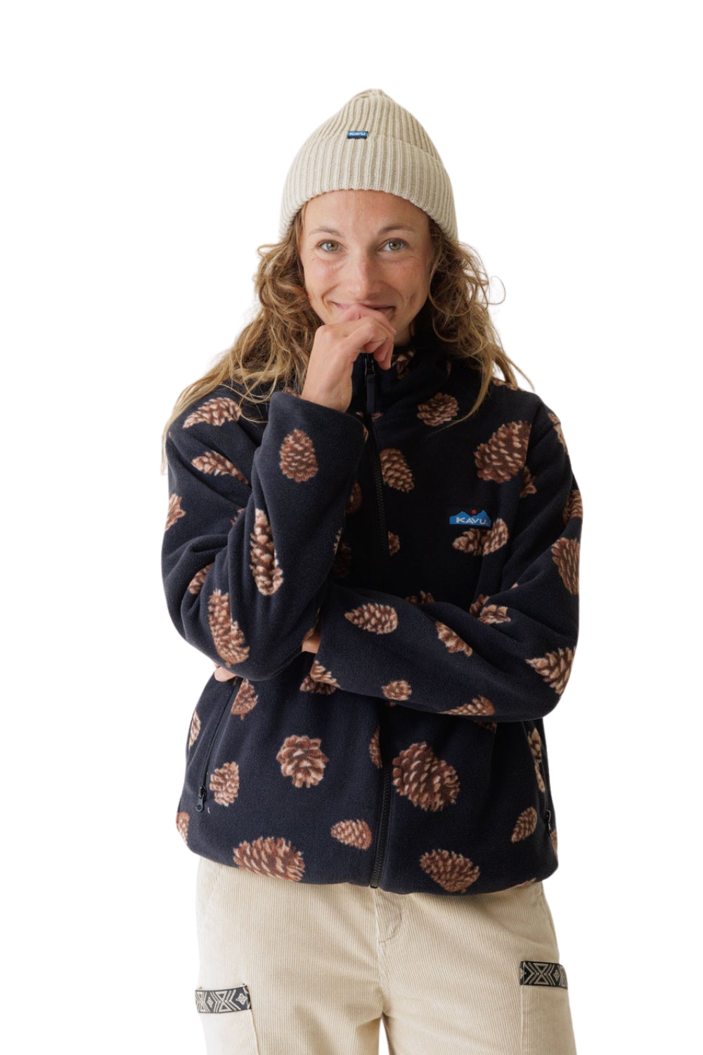 A woman wearing a navy Kavu fleece jacket with a brown pinecone pattern, a cream ribbed beanie, and beige pants, posing against a solid white background.