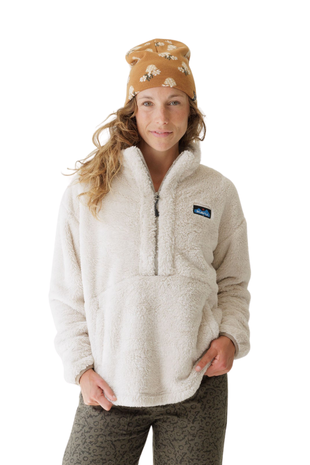 A woman wearing a cream-colored Kavu fleece pullover jacket with a half-zip, a tan patterned beanie, and patterned pants, photographed against a solid white background.