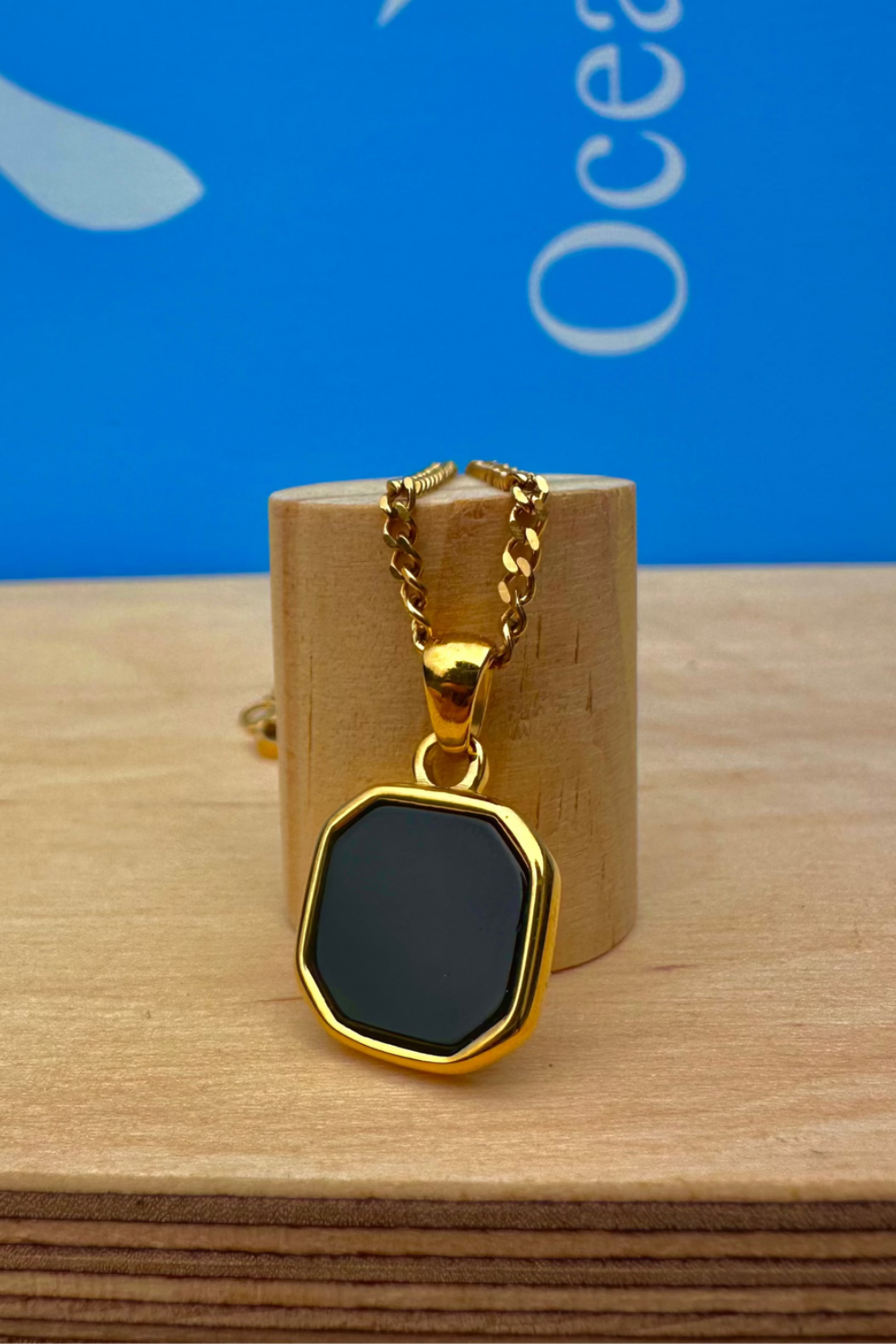 Gold necklace with a black pendant on a wooden stand against a blue background
