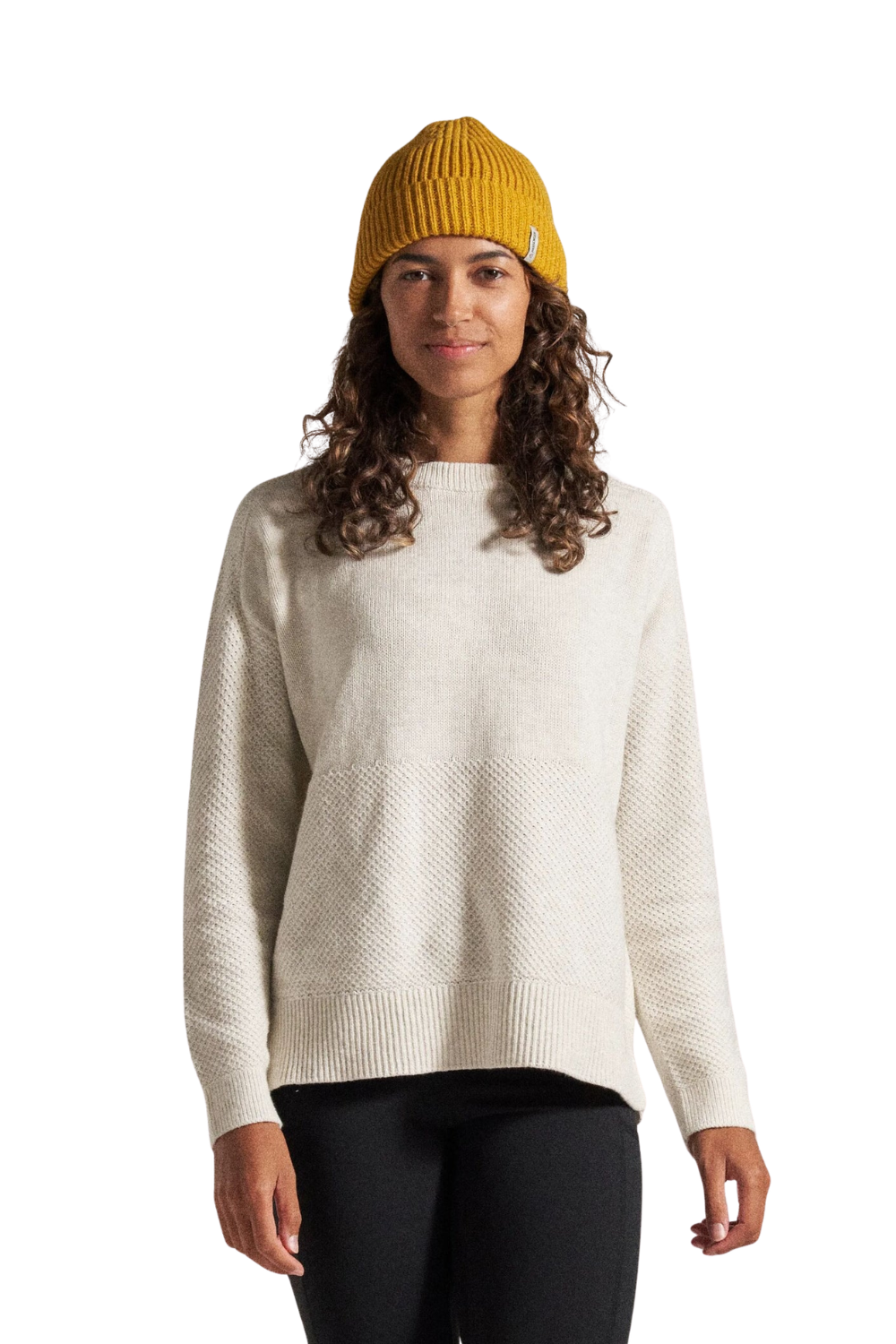 A woman wearing a cream-colored textured knit sweater, black pants, and a mustard yellow beanie, standing against a solid white background.