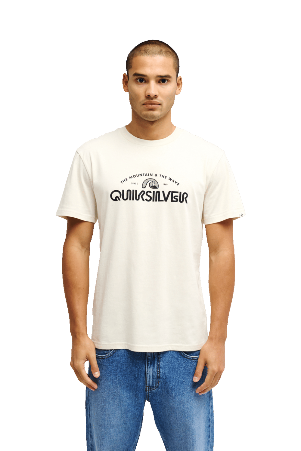 A man wearing a cream-colored Quiksilver t-shirt with a black graphic logo on the chest, paired with blue jeans, standing against a plain white background.