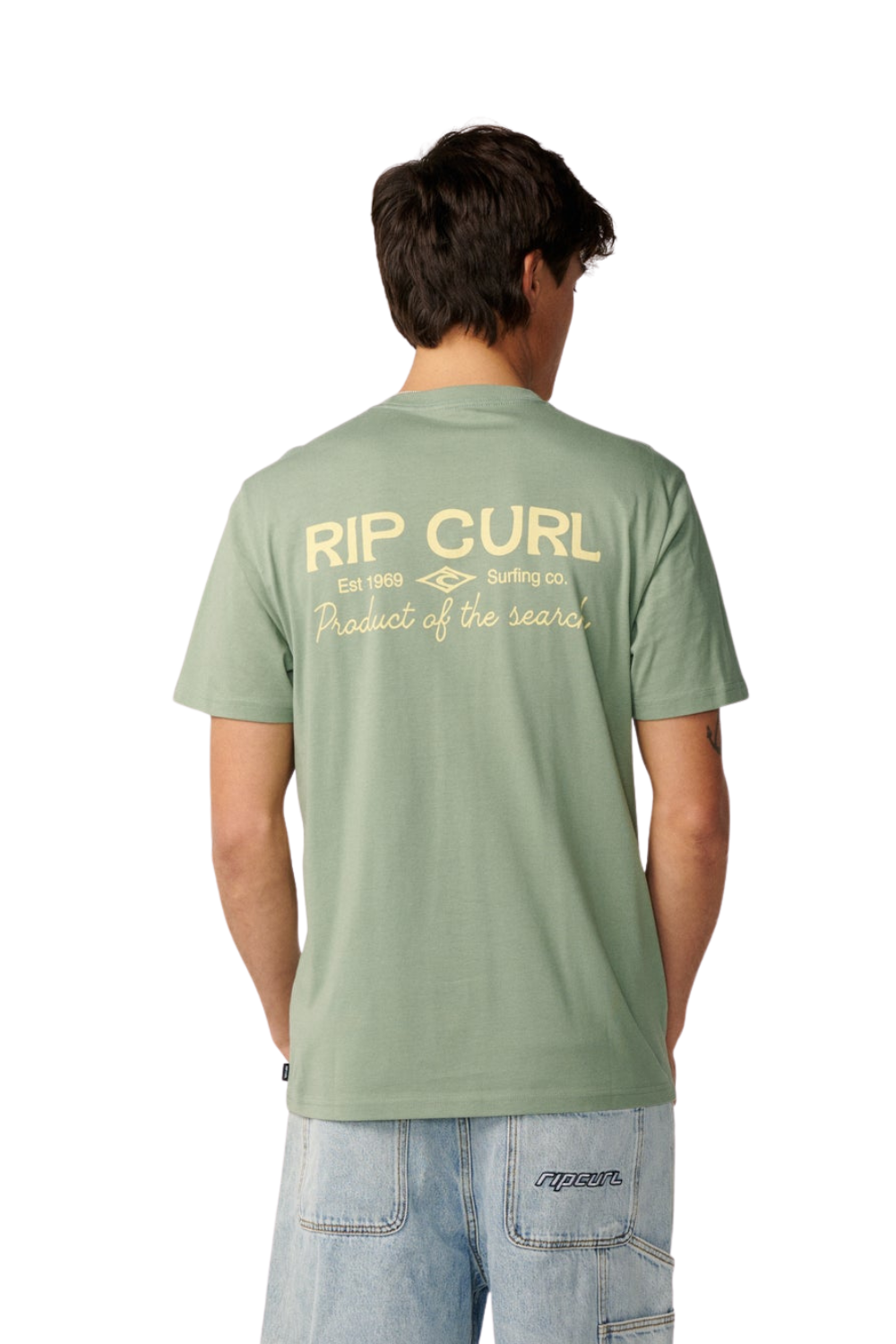 A man viewed from behind wearing a sage green Rip Curl t-shirt with a cream-colored graphic on the back, paired with light-wash jeans, against a plain white background.
