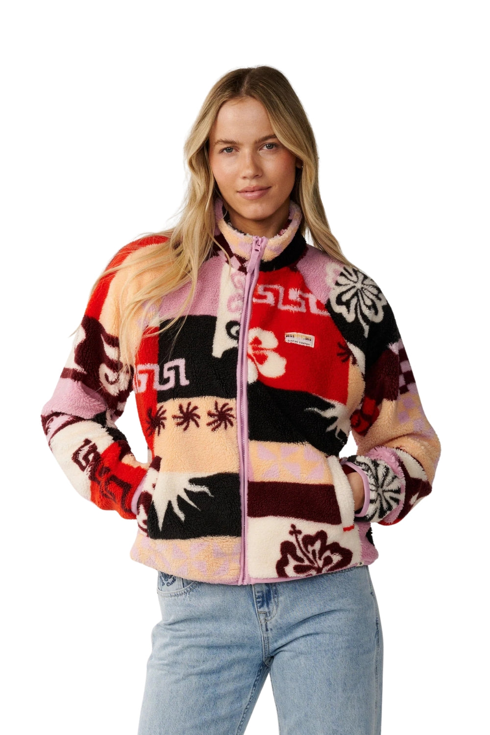 A woman wearing a colorful patchwork fleece zip-up jacket with Hawaiian and tribal-inspired patterns in red, pink, black, and cream, paired with light blue jeans, against a plain white background.
