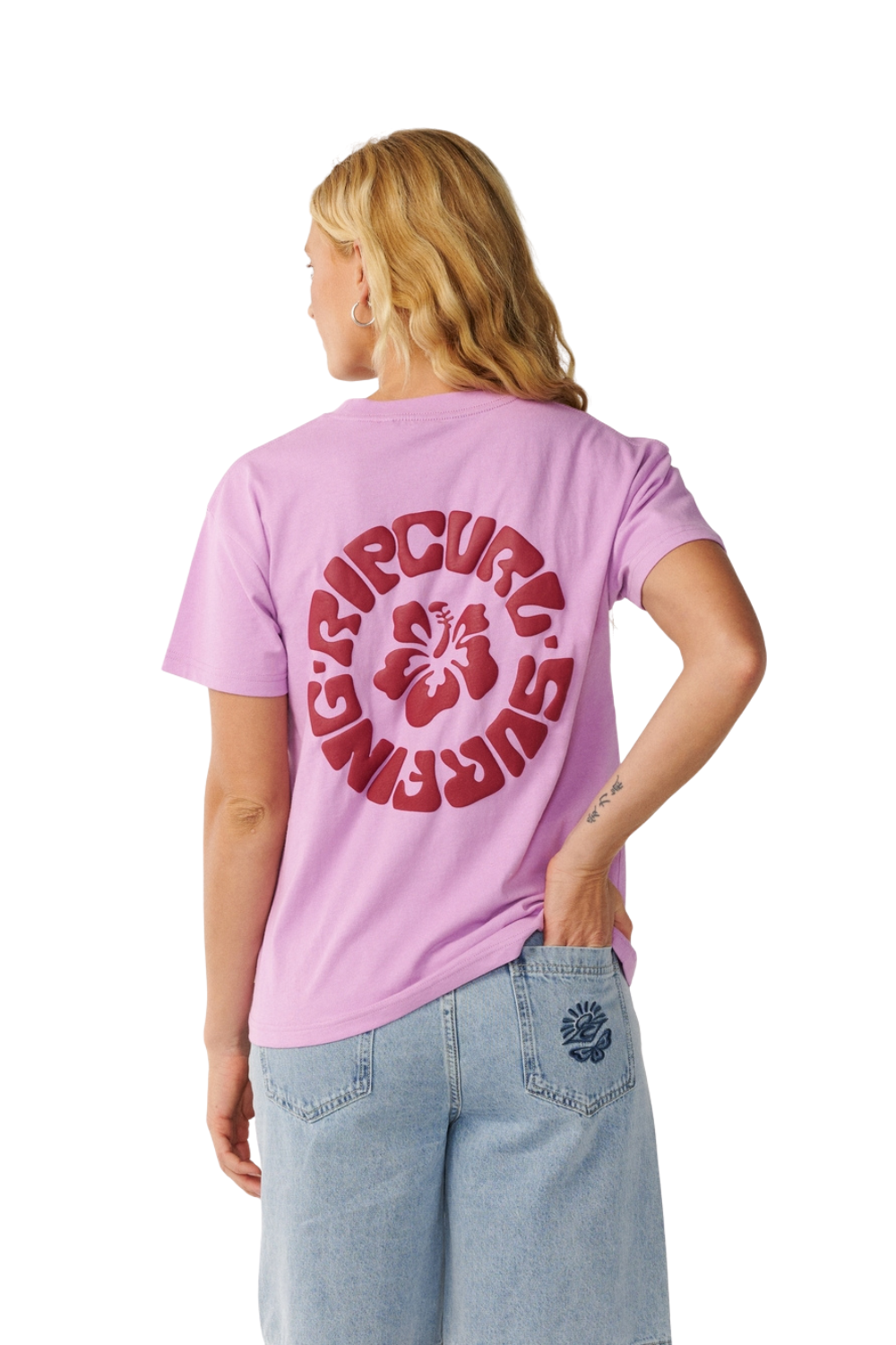 A woman shown from the back wearing a light purple t-shirt with a large red circular 'Rip Curl Surfing' graphic and hibiscus flower on the back, paired with light blue jeans against a white background.