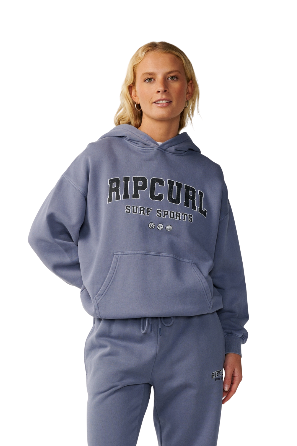 A woman modeling a dusty blue pullover hoodie and matching sweatpants set. The hoodie features 'RIP CURL SURF SPORTS' printed in dark gray across the chest, and the matching sweatpants have a small logo on the left thigh.