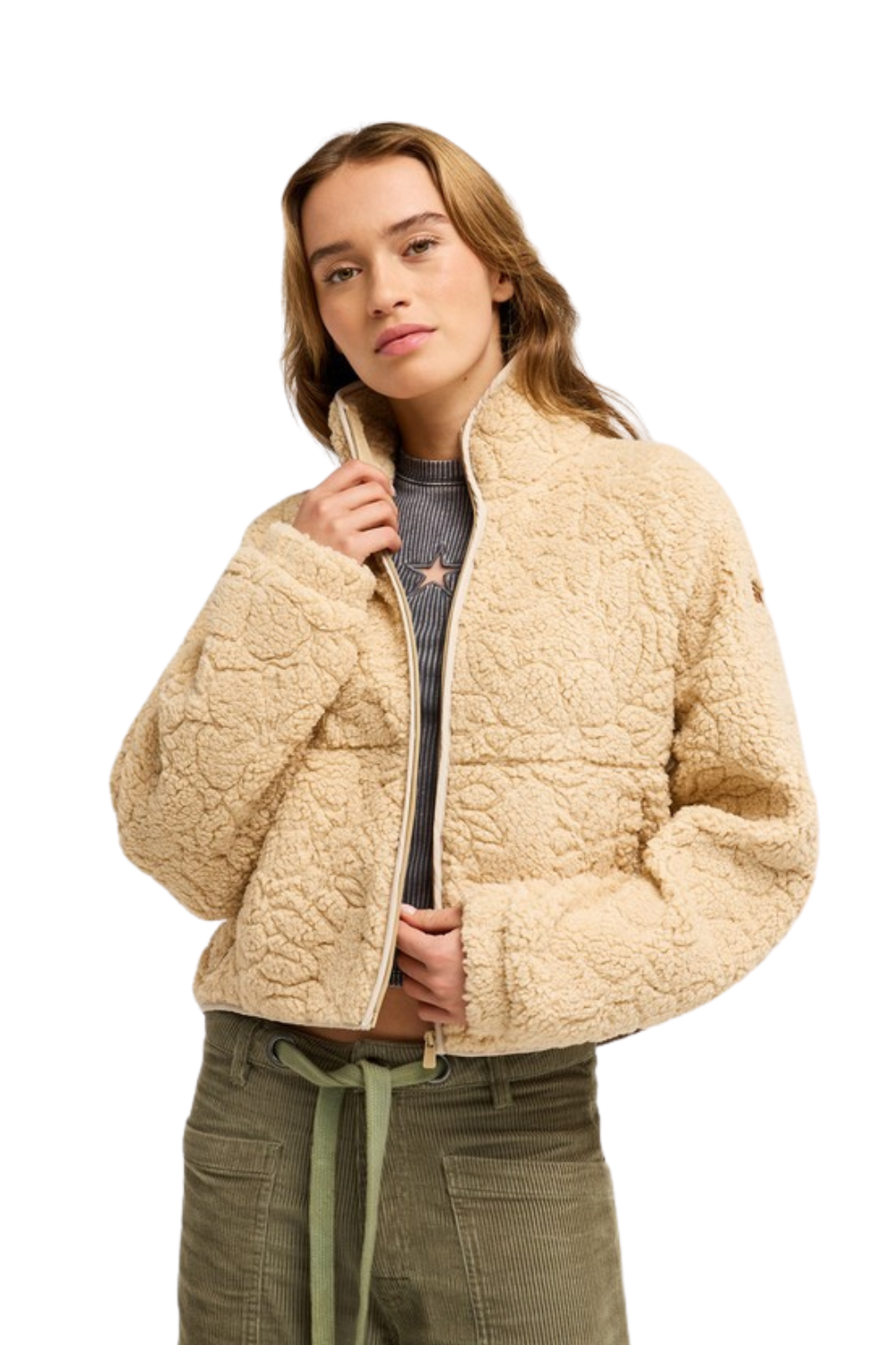 A woman modeling a cropped, quilted sherpa fleece jacket in a light beige color, paired with olive green pants and a gray top, against a solid white background.