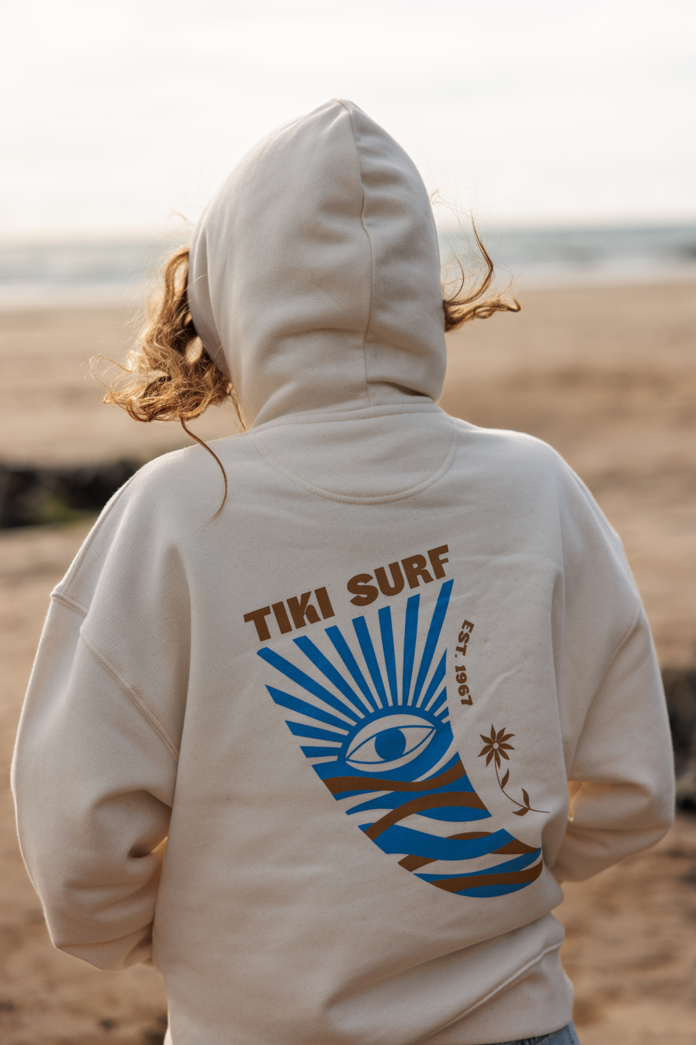 A person standing on a beach, viewed from behind, wearing a cream-colored hoodie with a large graphic on the back. The graphic features the text 'TIKI SURF' in brown above a blue and brown sunburst eye design.