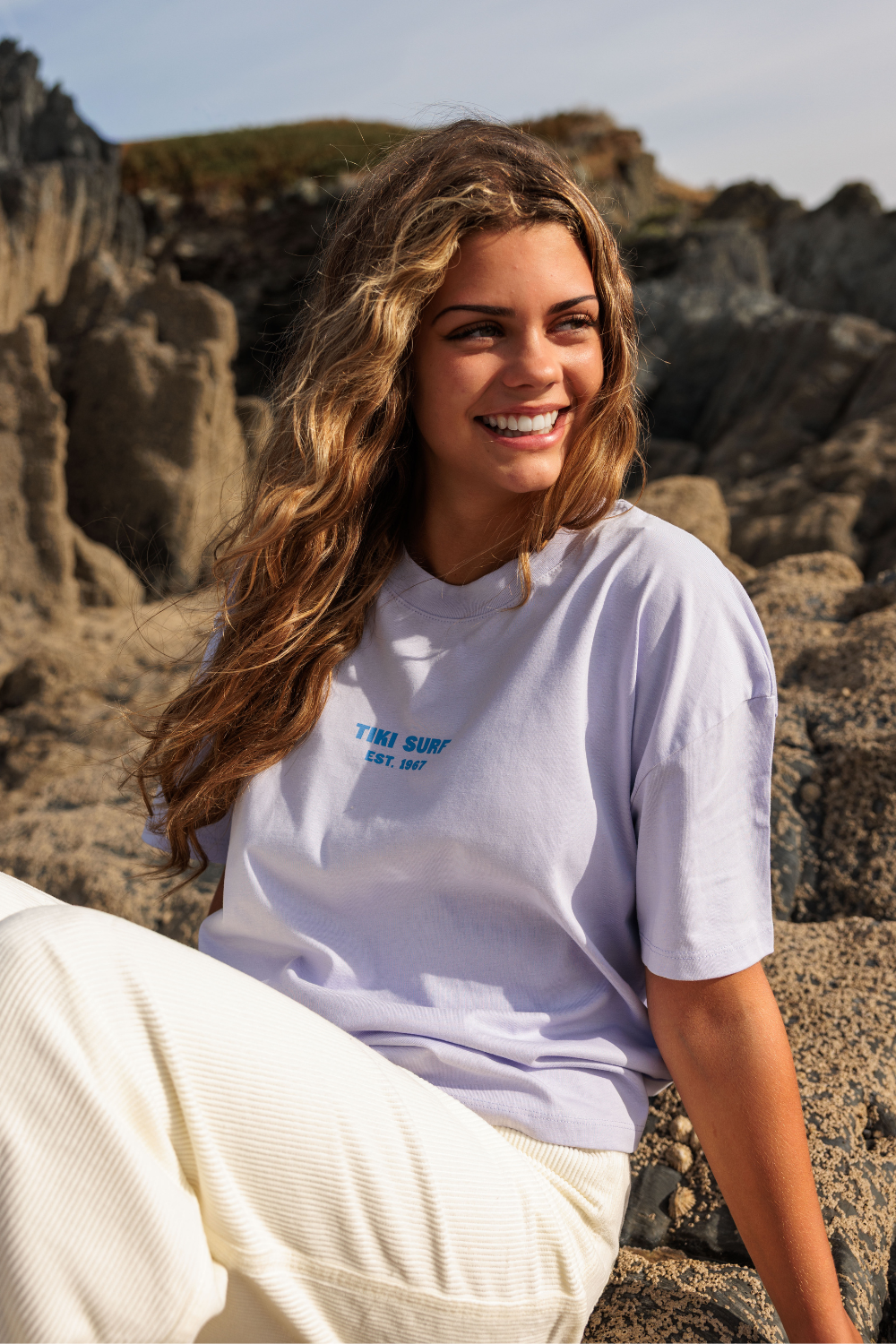 A woman with long wavy brown hair sits outdoors on rocky terrain wearing a light lavender t-shirt with blue text that reads 'THE SURF EST. 2017' and white pants.