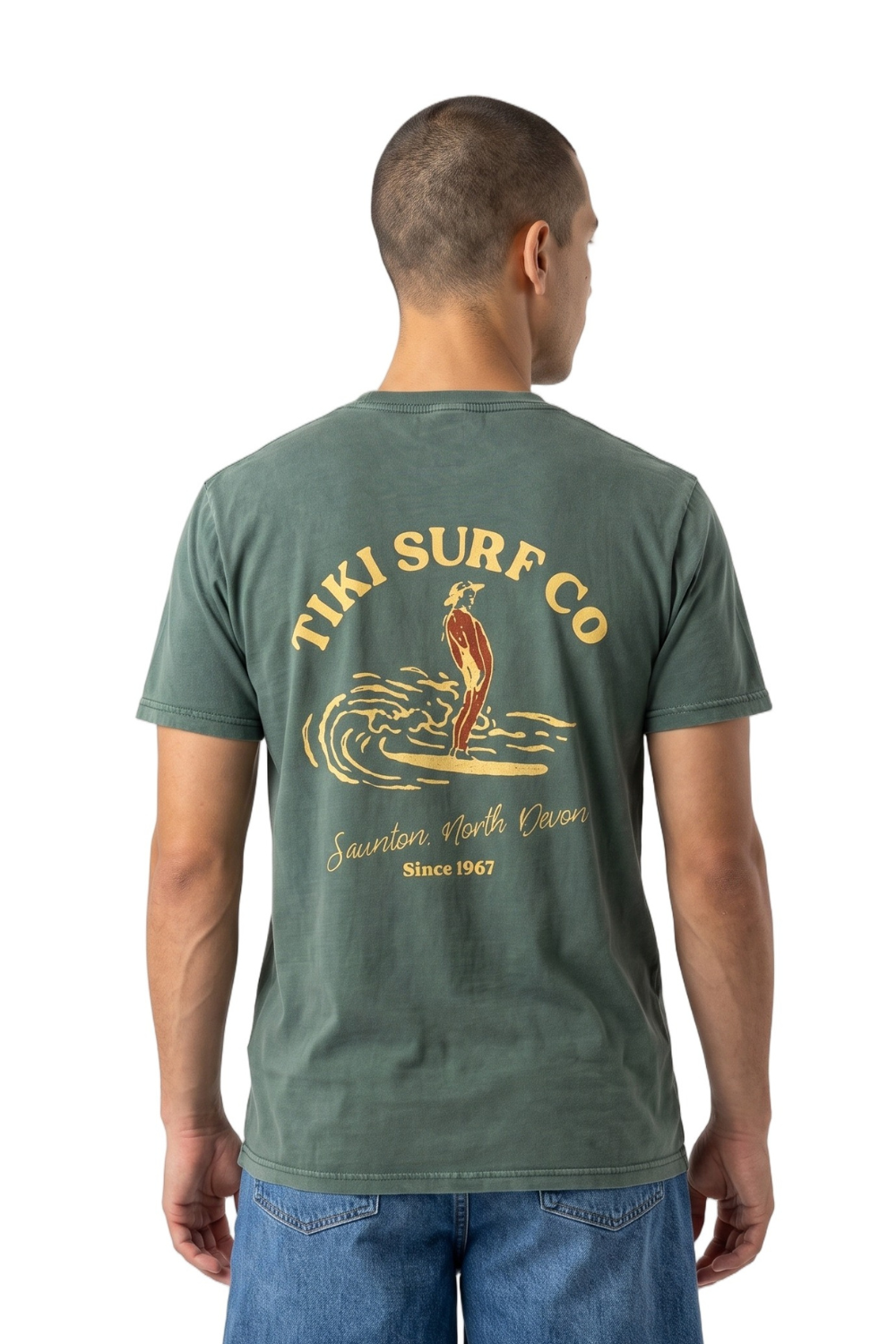 A man viewed from the back wearing a muted green T-shirt with a large vintage-style 'Tiki Surf Co' graphic on the back, paired with blue jeans, against a plain white background.