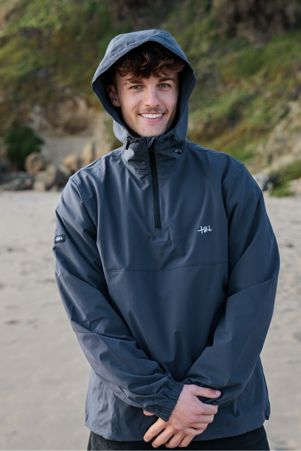 A man wearing a dark slate blue hooded quarter-zip anorak jacket, standing on a sandy beach with a rocky, grassy hill in the background.