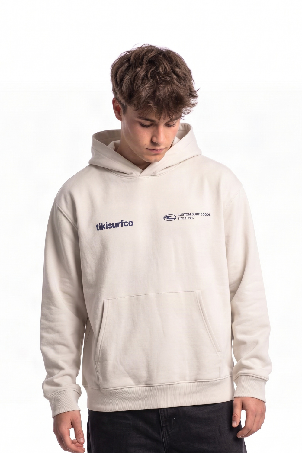 A young man wearing a cream-colored pullover hoodie with 'tikisurfco' printed in navy blue on the left chest, photographed against a solid white background.