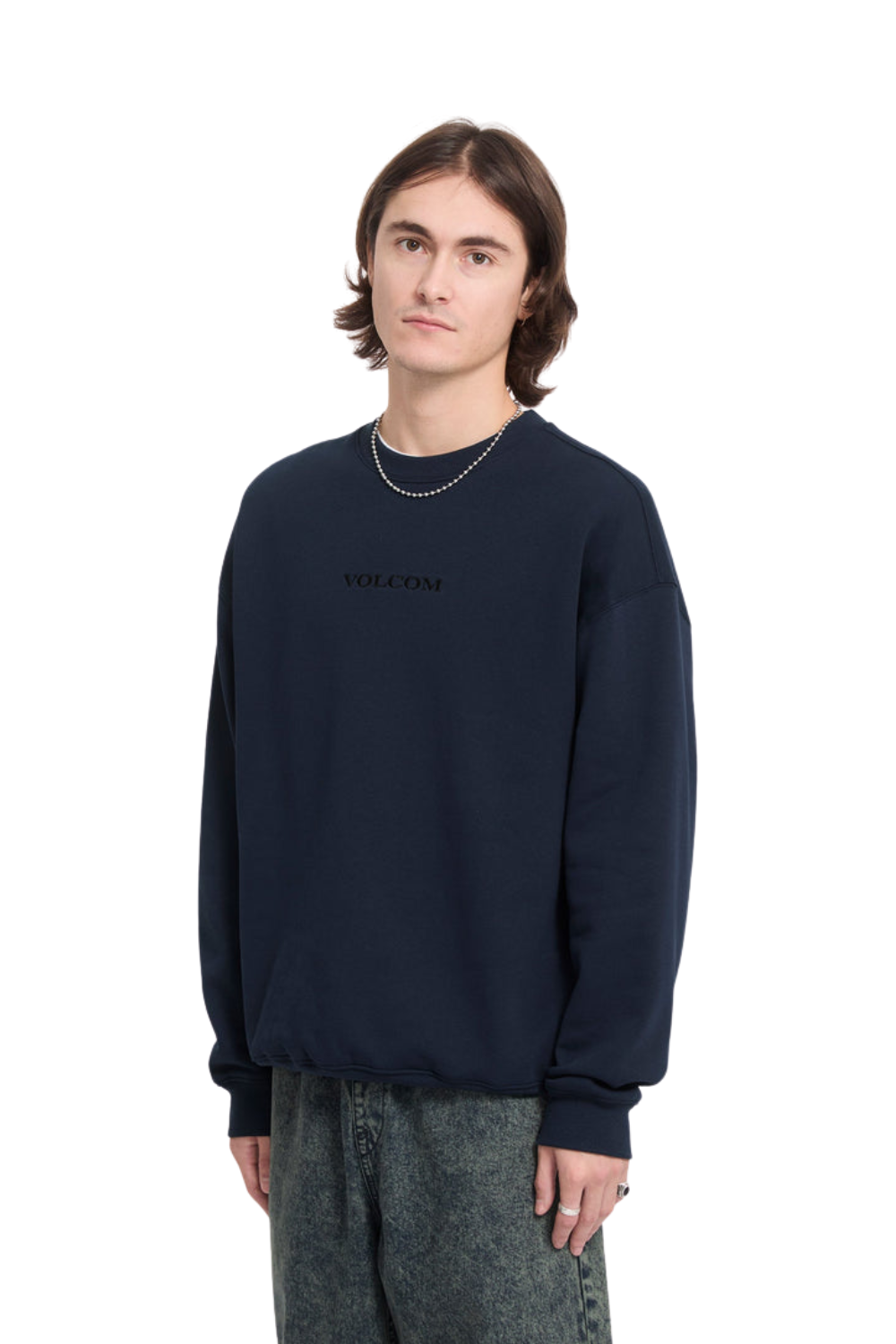 A young man wearing a dark navy blue crewneck sweatshirt with a subtle black 'Volcom' logo on the chest, paired with gray patterned pants, shot against a solid white background.