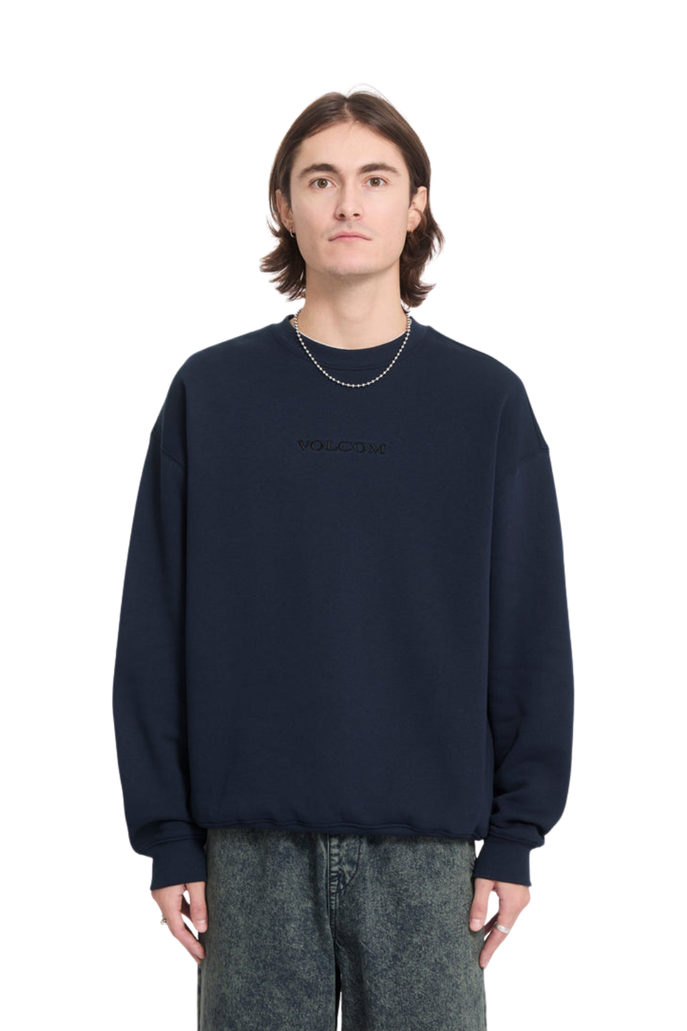 A man wearing a dark navy blue crewneck sweatshirt with a subtle tonal embroidered Volcom logo on the chest, paired with gray textured jeans and a silver chain necklace, shot against a plain white background.