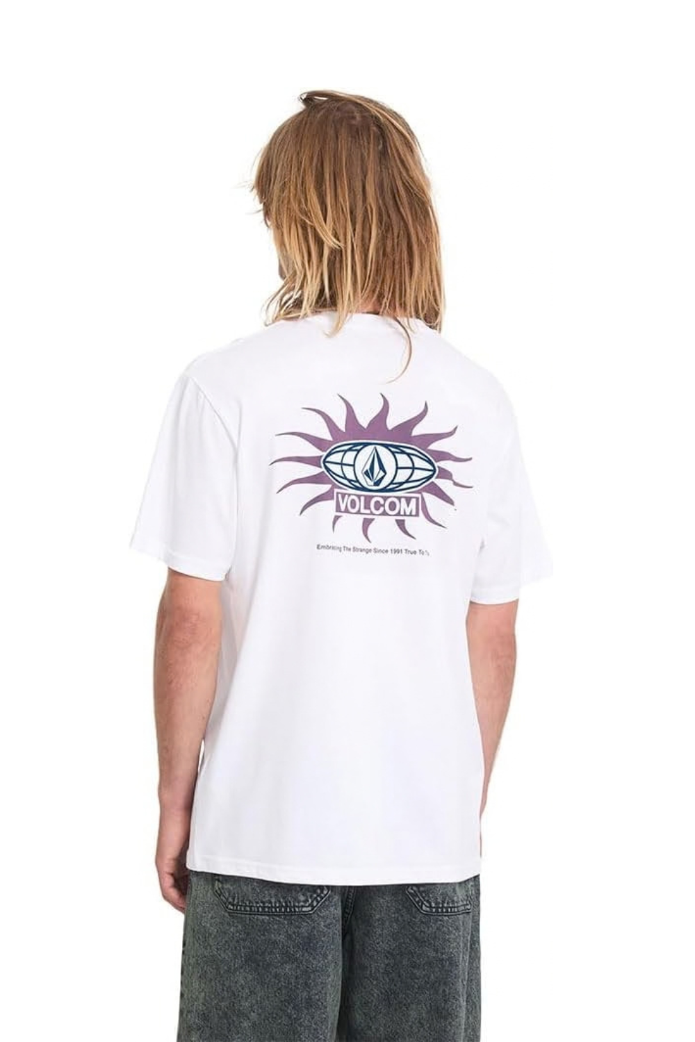 A model from behind wearing a white Volcom t-shirt with a purple and blue sunburst graphic on the back, paired with gray denim shorts against a white background.
