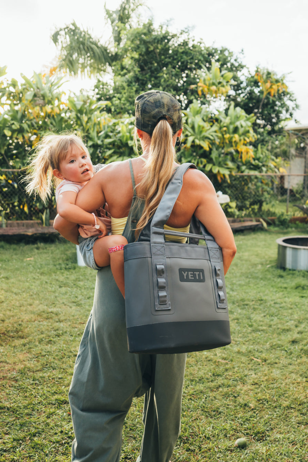 A woman outdoors carrying a young child on her back while wearing a large gray YETI tote bag over one shoulder. The scene is set in a grassy backyard with tropical foliage in the background.