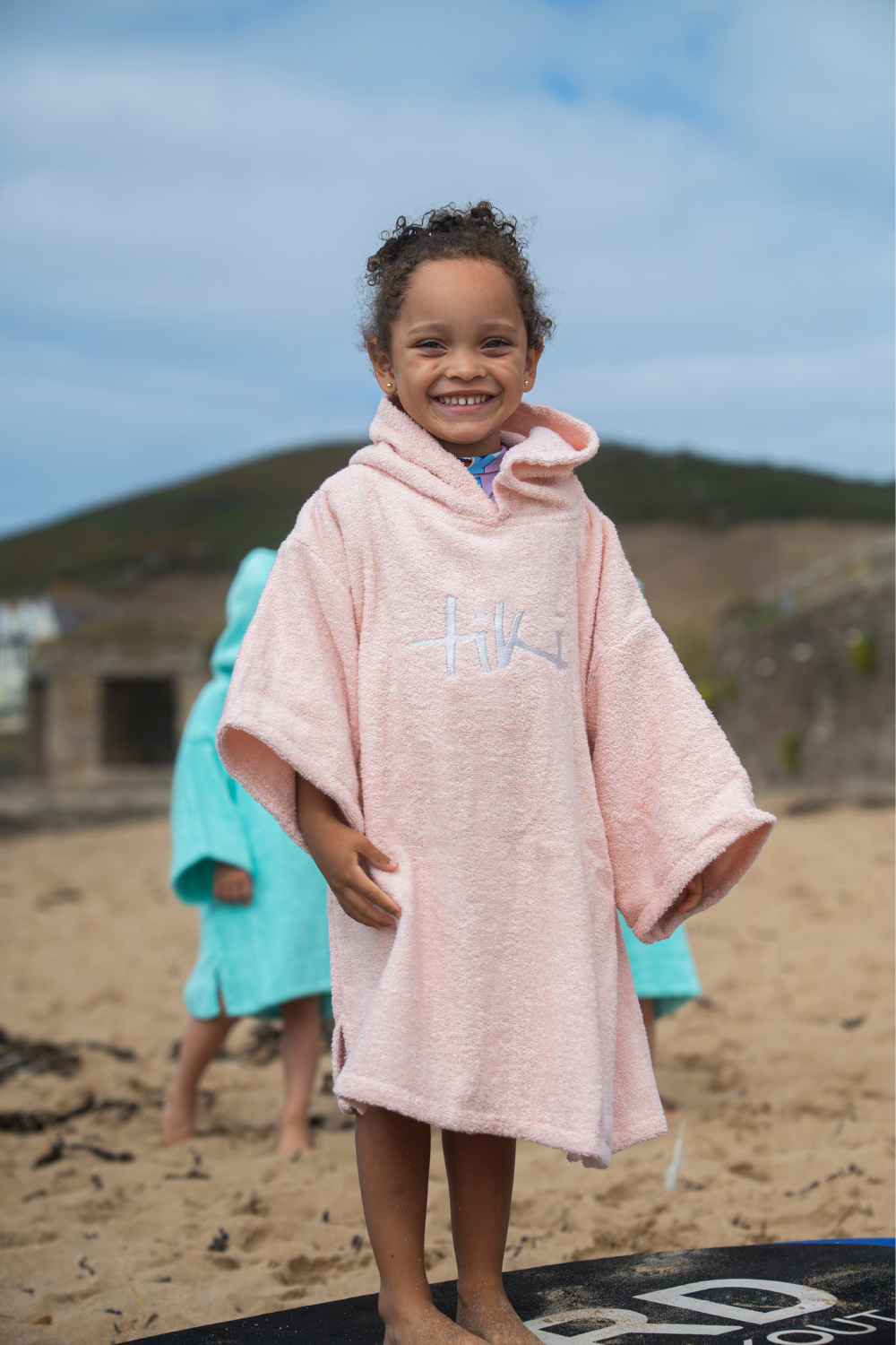 A smiling young child stands on a sandy beach wearing a light pink hooded towel poncho with 'Tiki' embroidered on the chest. 