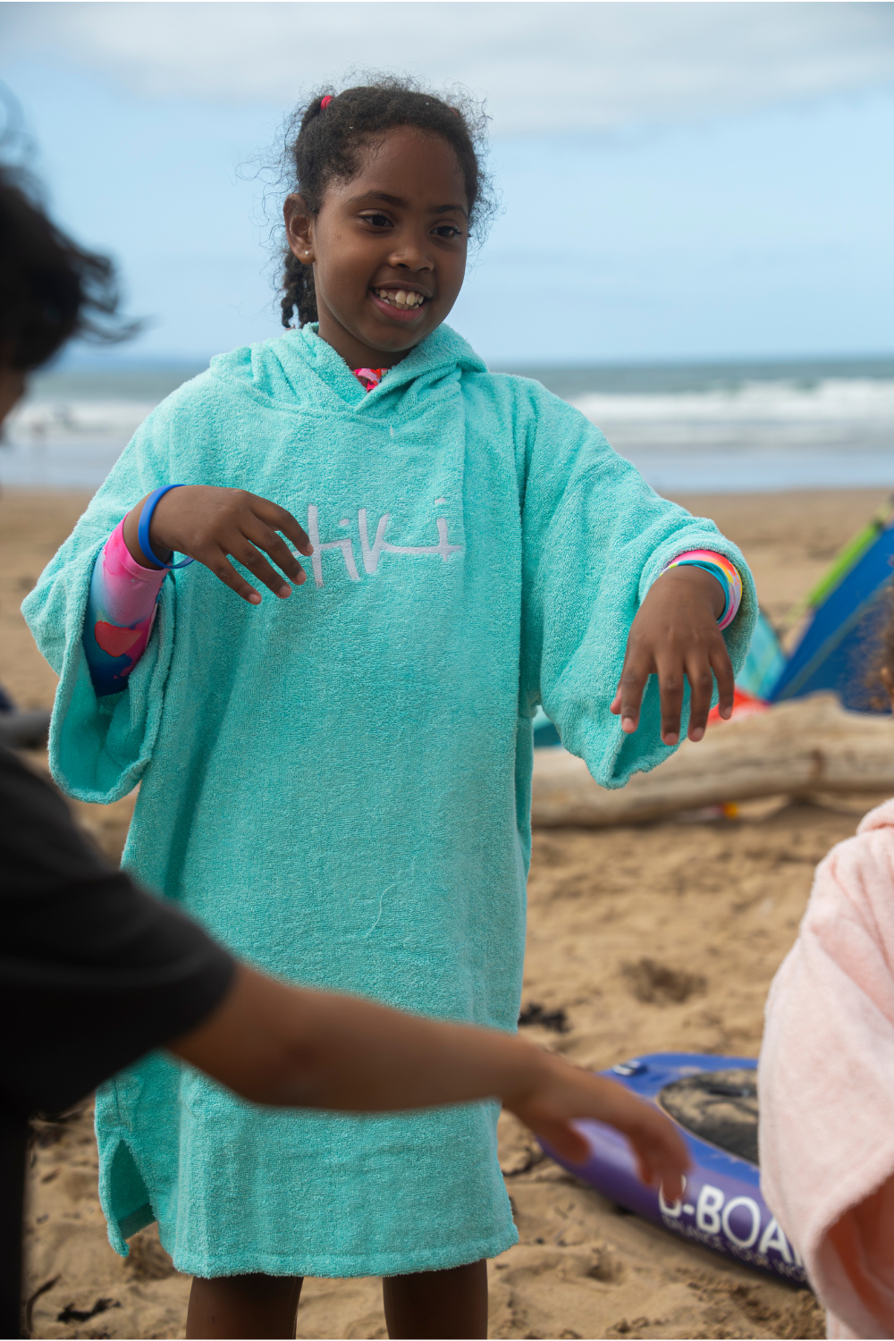 A young girl smiling on a sandy beach wearing a light aqua hooded towel poncho with 'Tiki' embroidered in white across the chest.