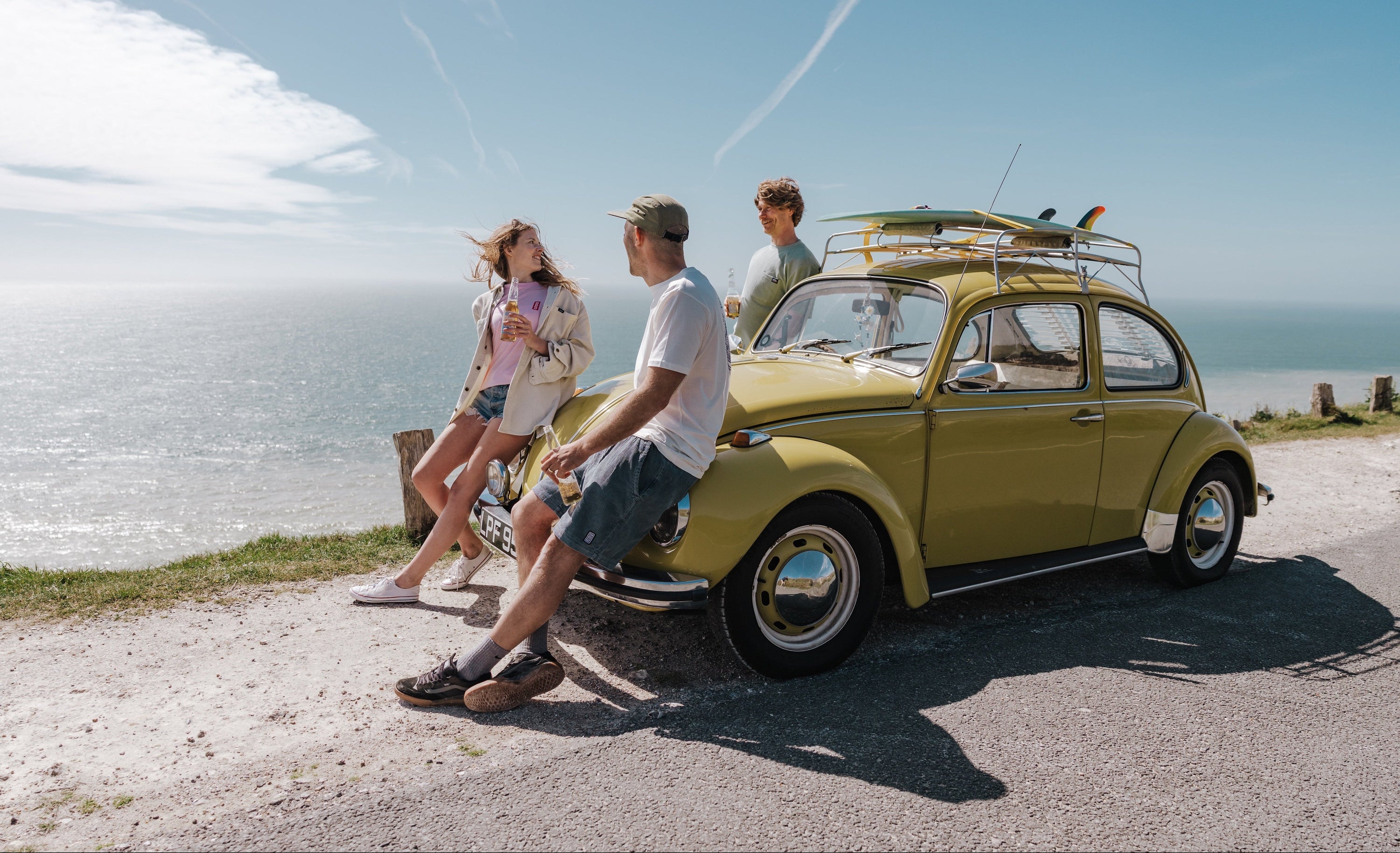 Three people by a vintage yellow car with a surfboard on a coastal road.