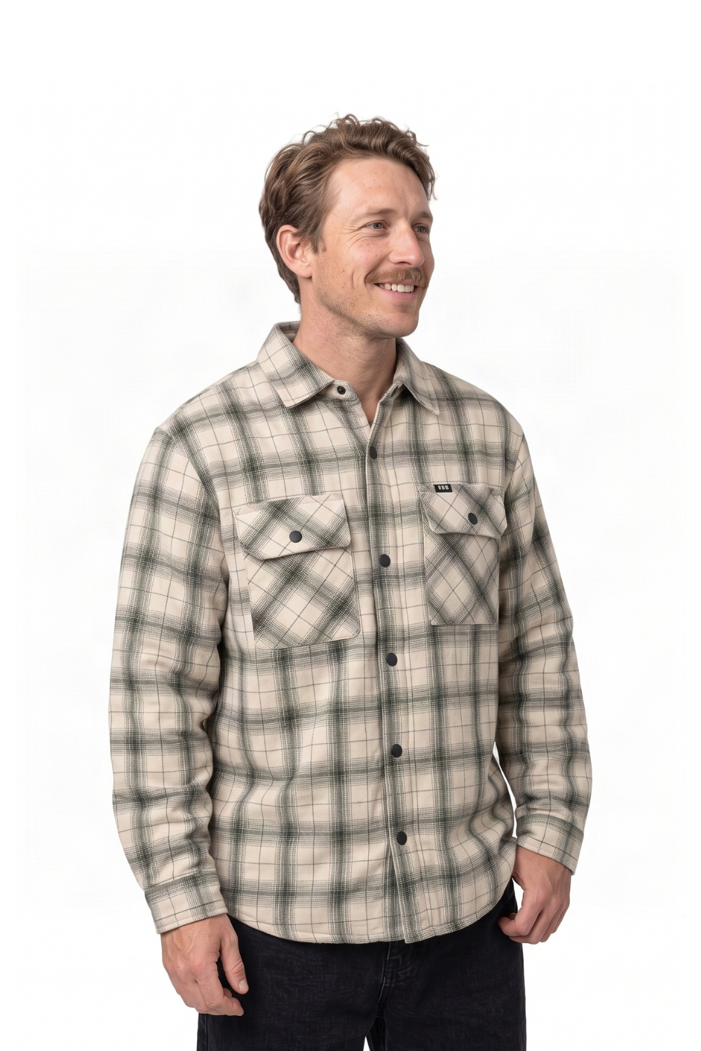 A man wearing a beige and gray plaid button-up flannel shirt, paired with dark jeans, standing against a plain white background.