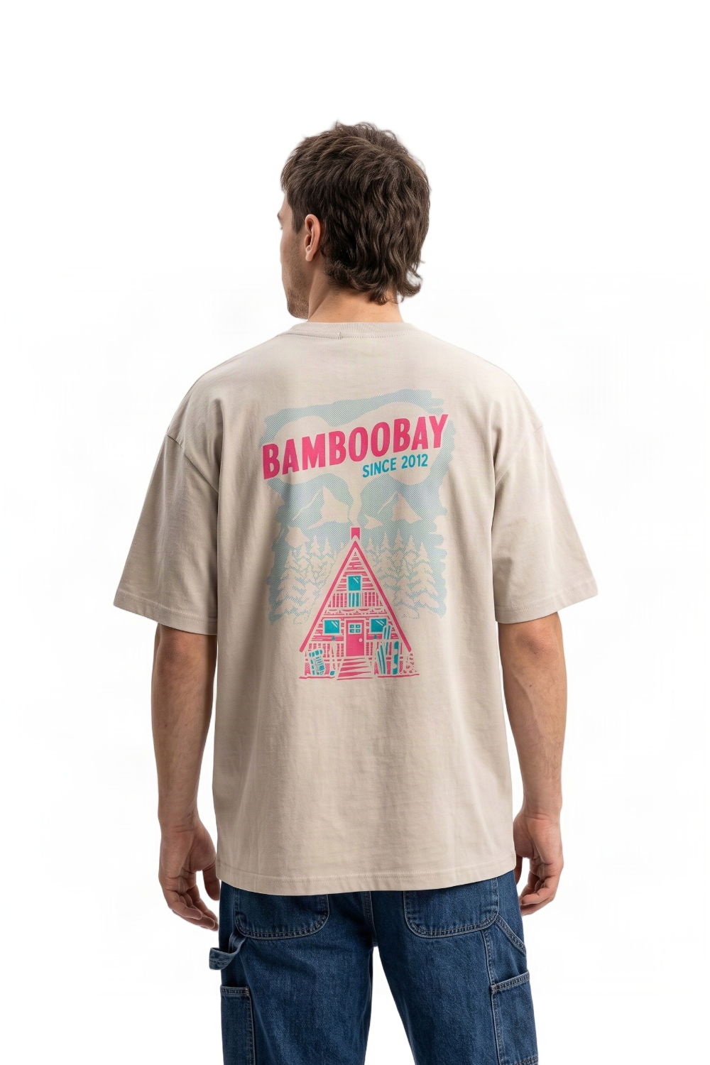 A man viewed from behind wearing a light beige t-shirt with a colorful graphic on the back that reads 'BAMBOOBAY SINCE 2022' above an illustration of a cabin and trees, paired with blue jeans against a white background.