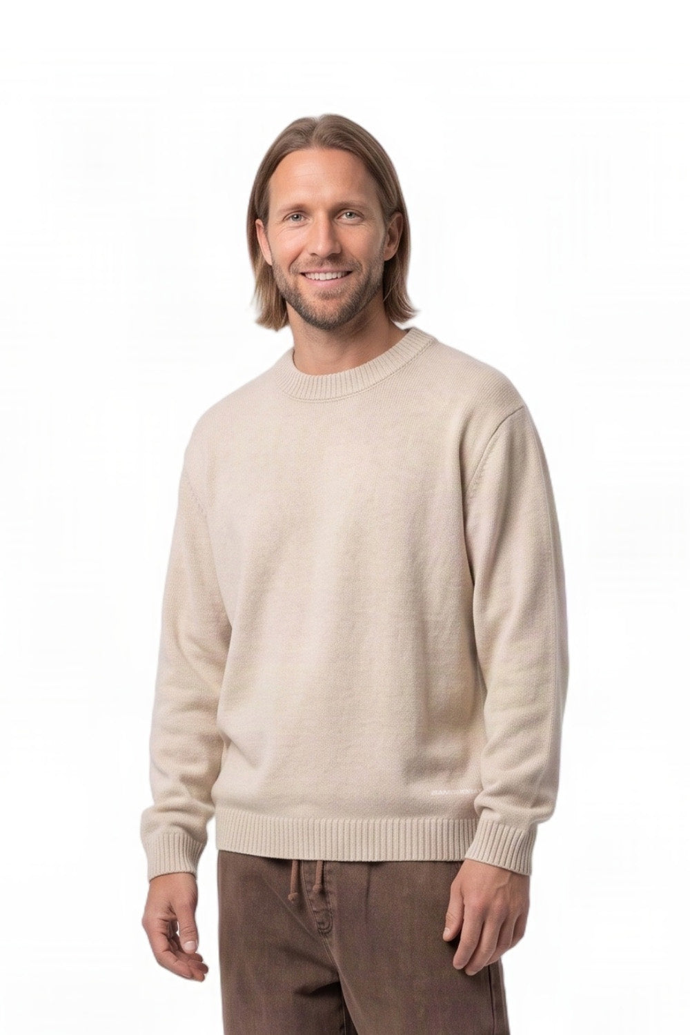A man with shoulder-length brown hair smiling, wearing a light beige crewneck knit sweater and brown pants, photographed against a solid white background.
