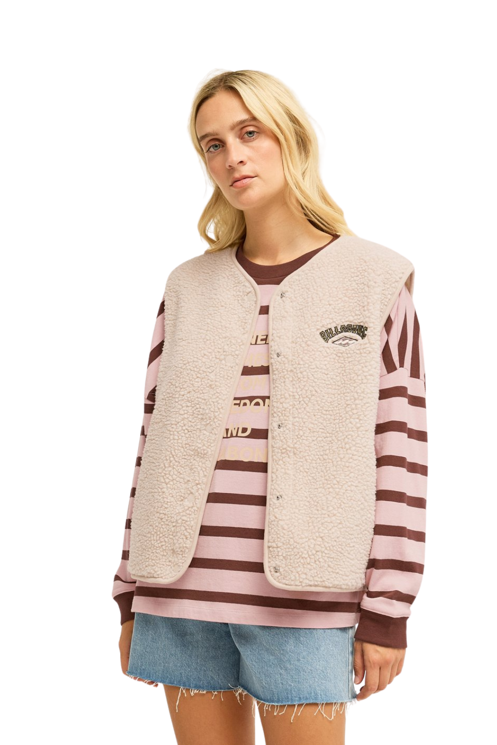 A blonde model wearing a beige Billabong sherpa fleece vest with pink and brown horizontal stripes on the sleeves and front, paired with denim shorts and a matching striped shirt.