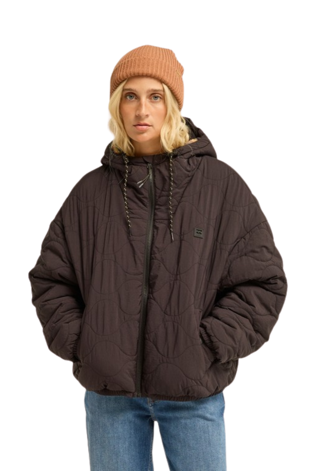 A model wearing a charcoal quilted puffer jacket with a hood, paired with a rust-colored beanie and blue jeans, against a plain white background.