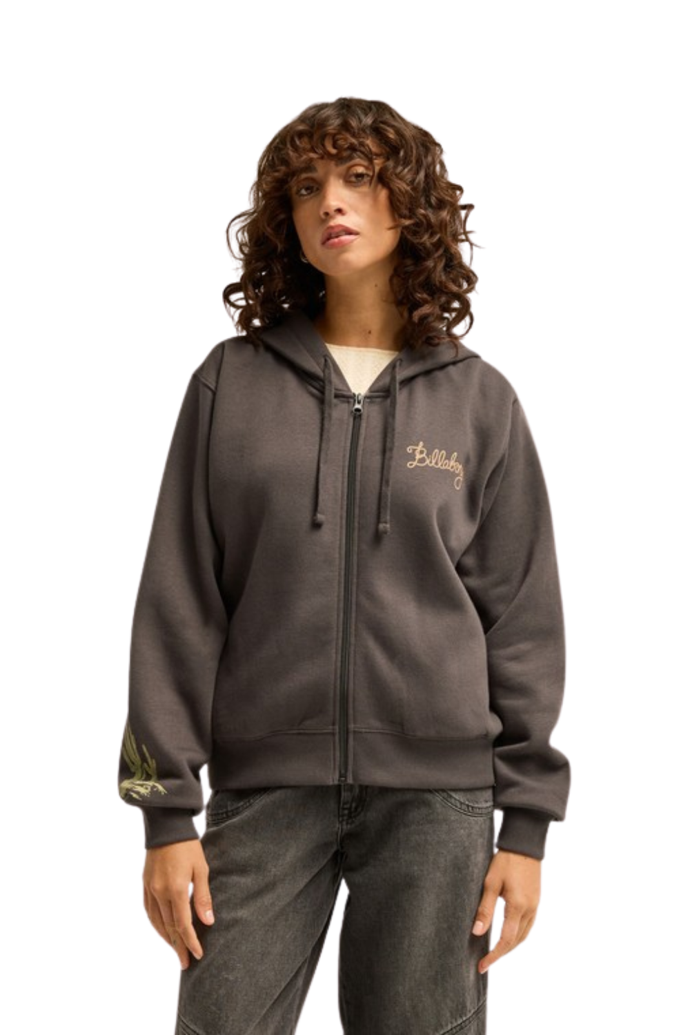 A woman wearing a charcoal zip-up hoodie with gold embroidered details on the chest and sleeve, paired with dark gray jeans, against a plain white background.