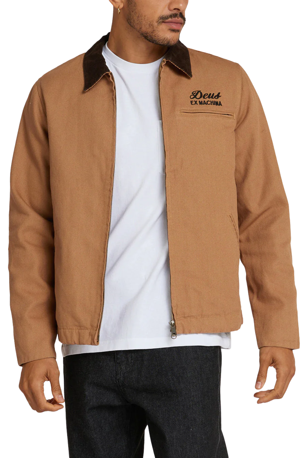 A man wearing a tan work jacket with a dark brown collar, layered over a white t-shirt and dark jeans. The jacket features a zippered front, a chest pocket with 'Deus' embroidered on it, and a front zipper.