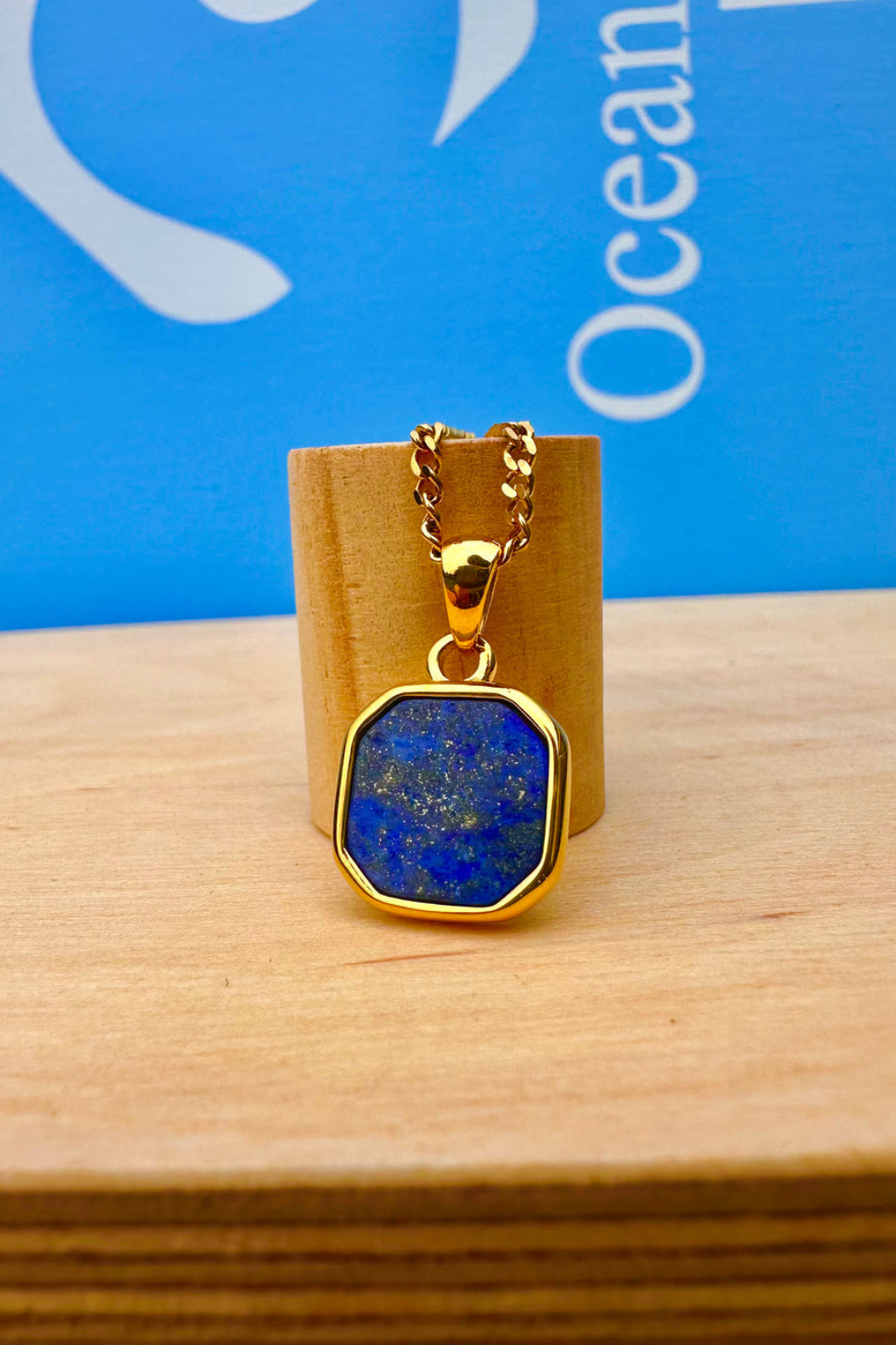 Gold necklace with blue lapis lazuli stone pendant on a wooden stand against a blue background