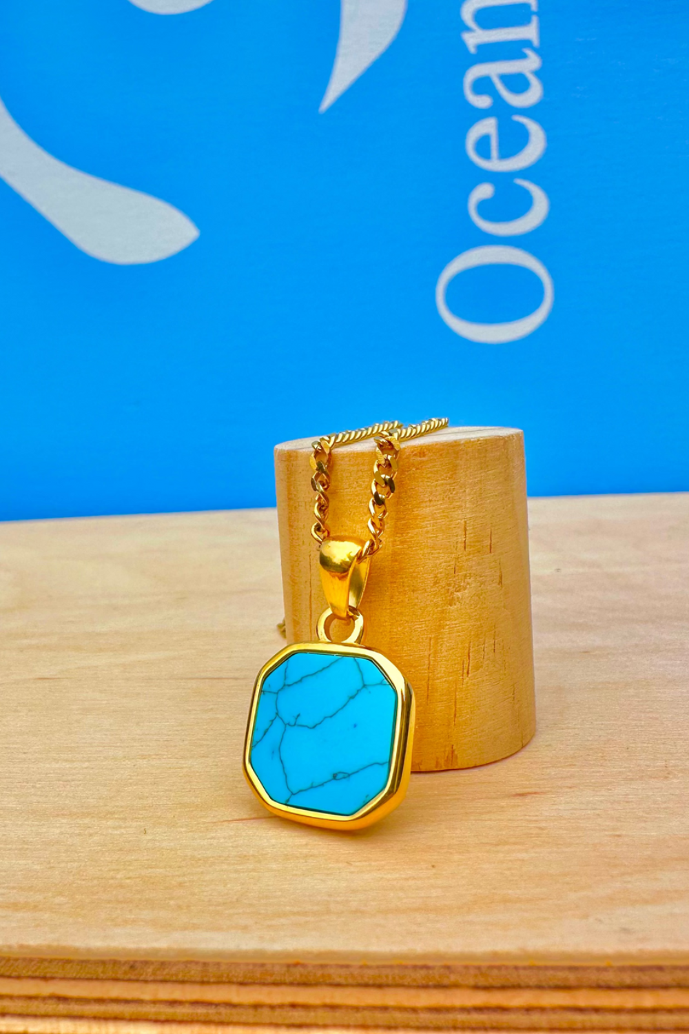 Gold necklace with a turquoise pendant on a wooden stand against a blue background.