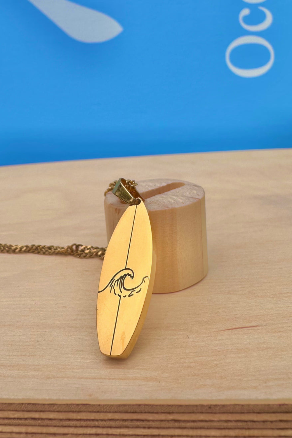 Gold necklace with a surfboard shaped and wave design pendant on a wooden surface