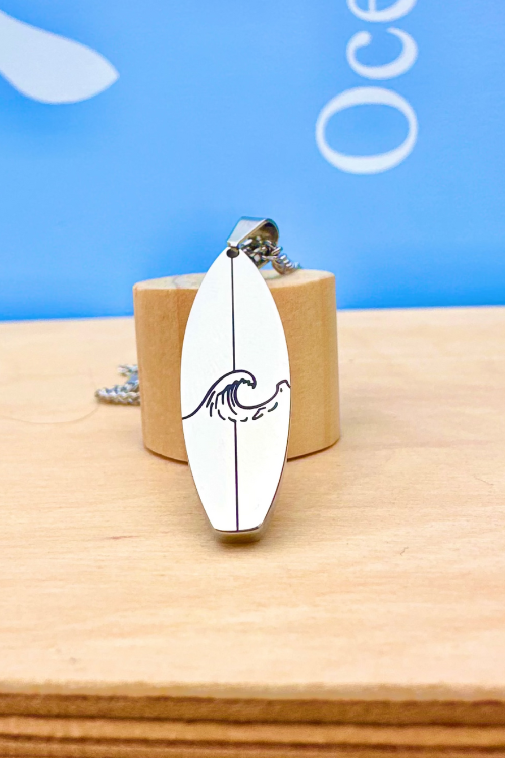 Surfboard-shaped pendant with wave design on a wooden block against a blue background 