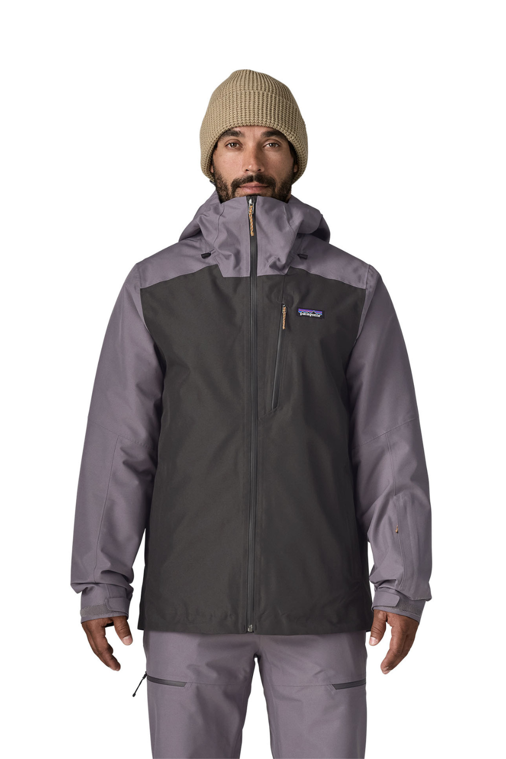 A man wearing a Patagonia hooded jacket in dark gray and muted purple-gray, paired with matching pants and a beige knit beanie, photographed against a plain white studio background.