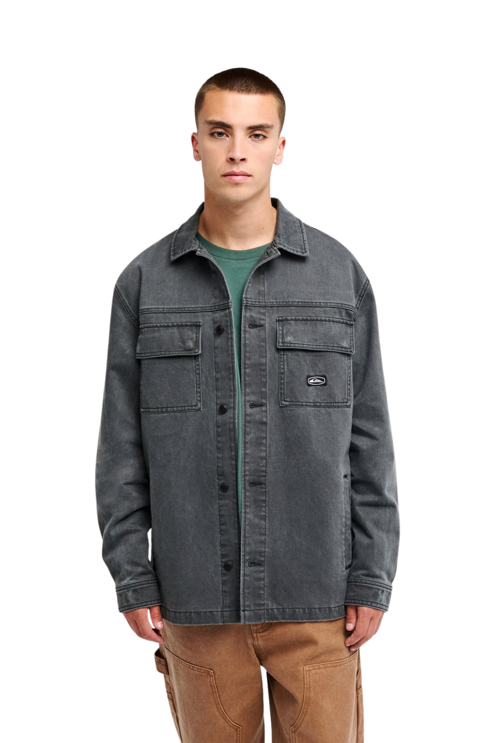 A man wearing a dark gray washed denim button-up shirt jacket over a green t-shirt and brown pants, standing against a solid white background.