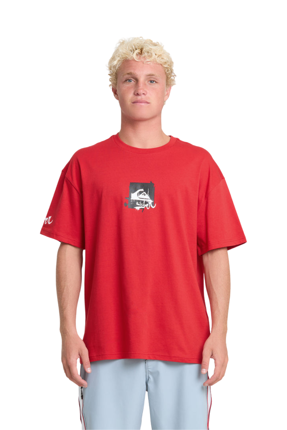 A blonde male model wearing a red Quiksilver t-shirt with a small black and white graphic on the chest, paired with light blue track pants, shot against a solid white background.