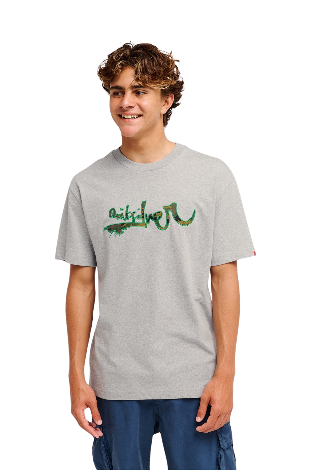 A smiling young man wearing a light heather gray t-shirt with a green and black graphic 'Quiksilver' logo across the chest, paired with dark blue cargo pants, set against a plain white background.