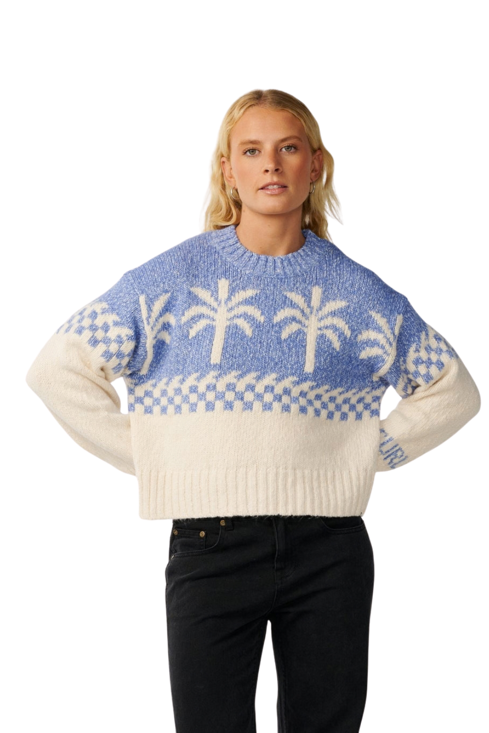 A woman modeling a knit sweater with a blue and cream checkerboard pattern, white palm tree motifs, and solid cream sleeves, paired with black jeans against a plain white background.