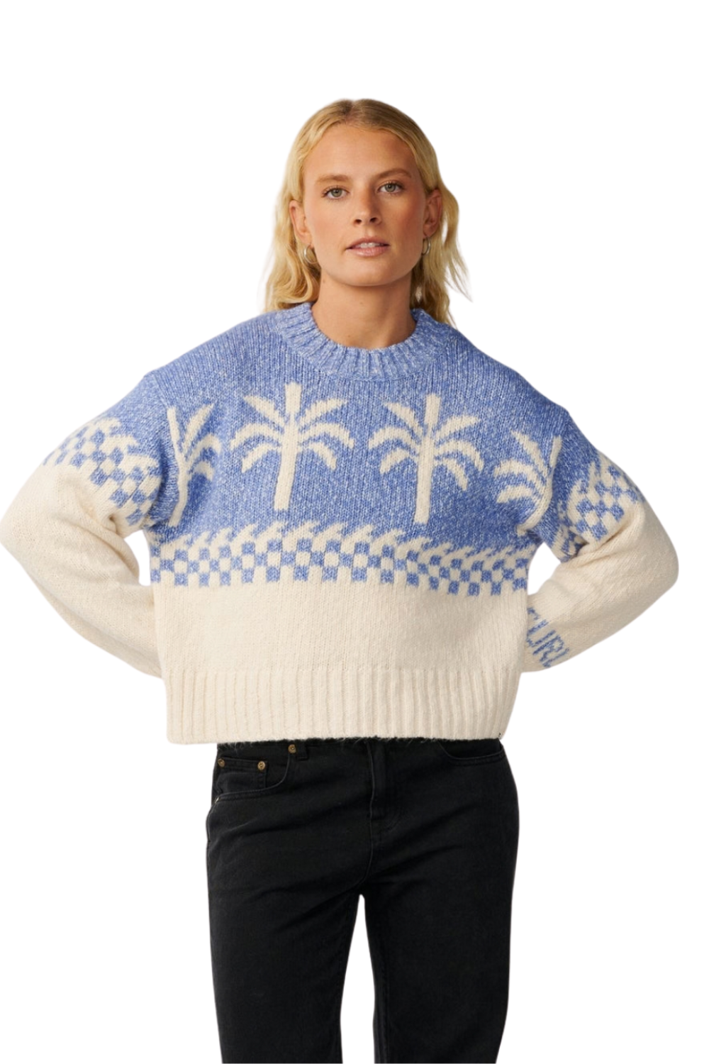 A woman modeling a light blue and cream knit sweater with palm tree and checkerboard patterns, paired with black jeans against a plain white background.