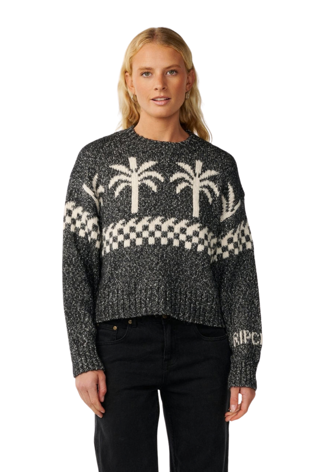 A woman modeling a dark gray and cream knit sweater with palm tree and checkerboard patterns, paired with black jeans against a plain white background.