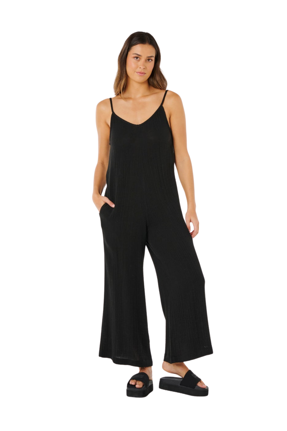 A woman modeling a black sleeveless jumpsuit with thin spaghetti straps and wide legs, paired with black slide sandals, against a plain white background.